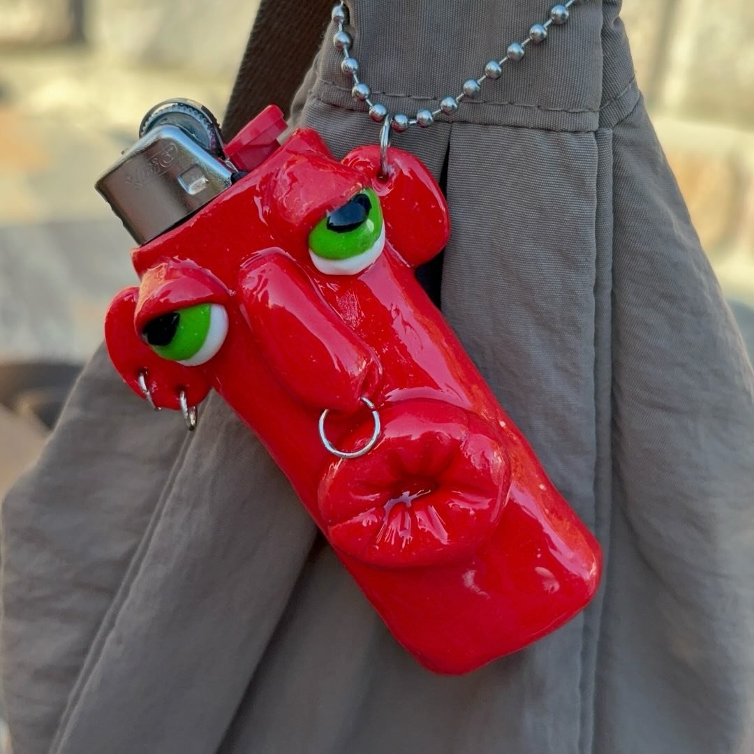 Handmodellierte Feuerzeug-Hülle in glänzendem Rot mit grünen Augen, Nasenring und Lippen, am Riemen einer Tasche befestigt